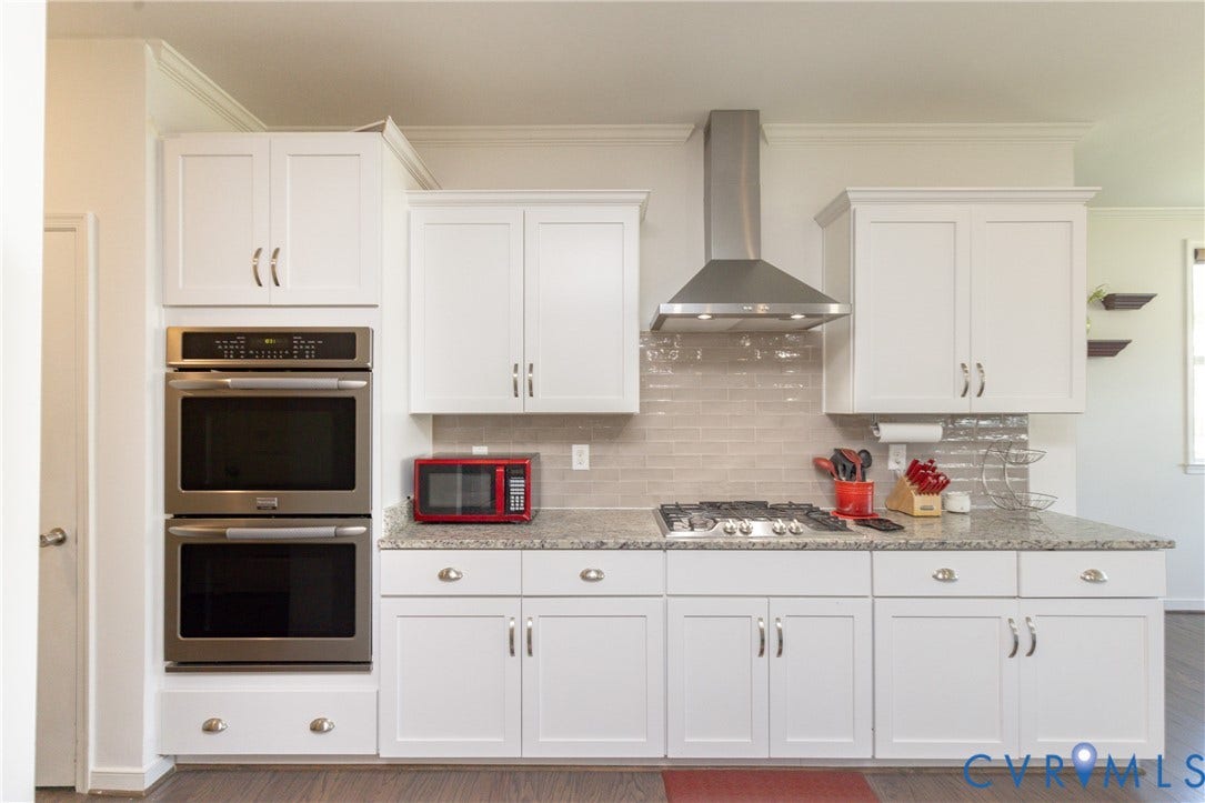 White shaker-style cabinetry, stainless steel double wall ovens, gas range with stainless steel hood, light-toned tile backsplash, and light-toned speckled countertops