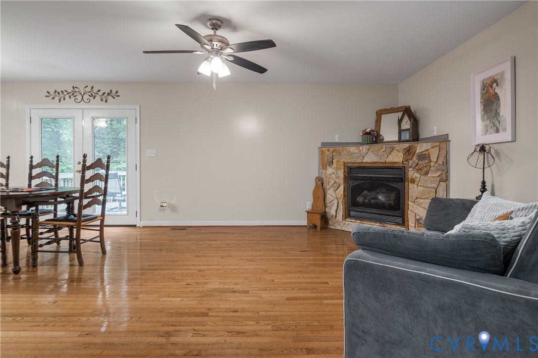 Hardwood flooring, natural stone fireplace surround, ceiling fan with integrated lighting, glass-paned French doors, and exterior deck access