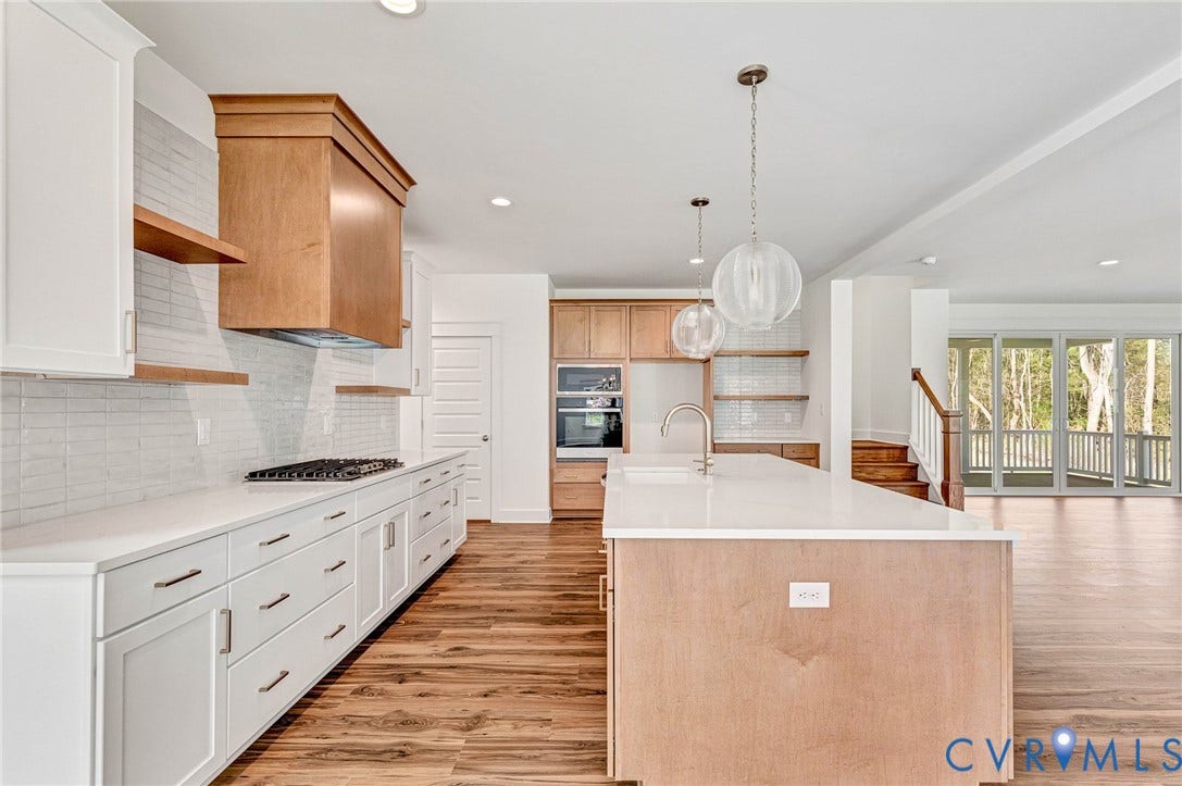 This kitchen features wood flooring, white cabinetry with dark hardware, a center island with a sink and pendant lighting, and a white tile backsplash