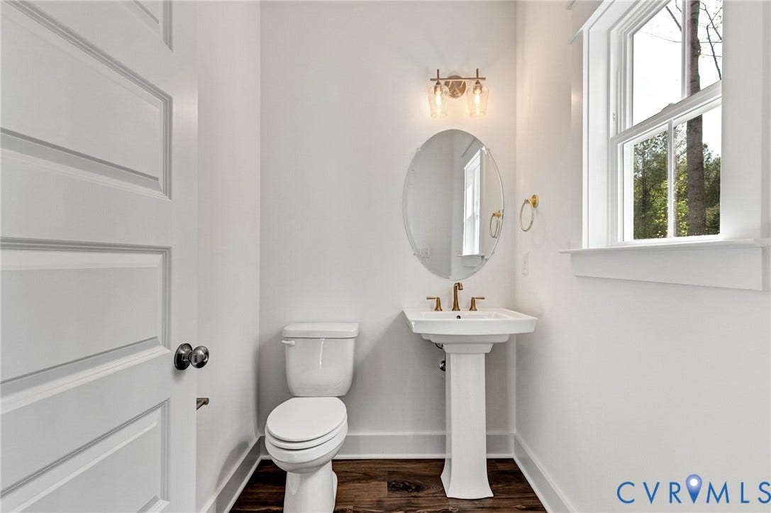 The bathroom features a pedestal sink with a gold-toned faucet, an oval mirror, and a window providing natural light