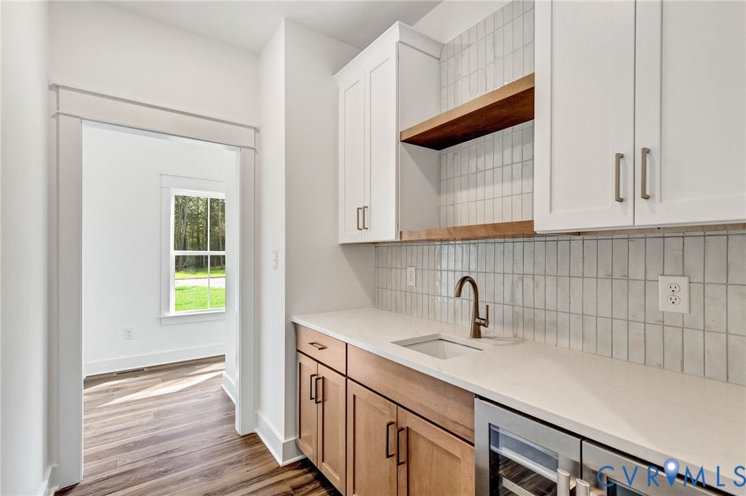 Wet bar area featuring white upper cabinetry, a wood shelf, a white tile backsplash, a sink with a bronze faucet, and light wood lower cabinetry with a built-in beverage cooler