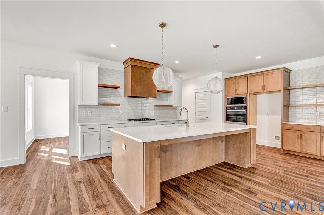 The kitchen features wood flooring, a central island with a sink, white cabinetry with open shelving, and integrated wall ovens