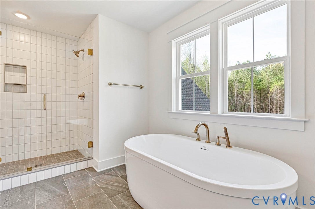A spacious bathroom featuring a freestanding bathtub with bronze fixtures, a glass-enclosed shower with white tiled walls, and large windows providing natural light and views of trees