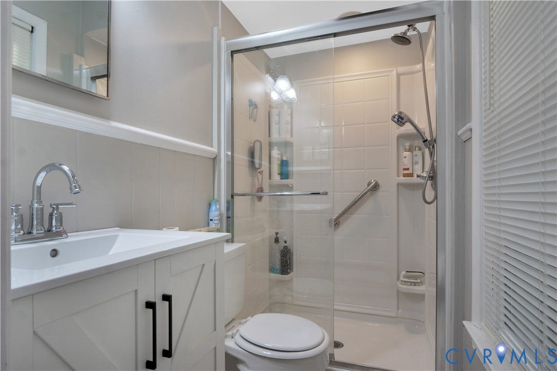 White vanity with black hardware, chrome faucet, white sink, and glass-enclosed shower with a handheld shower head
