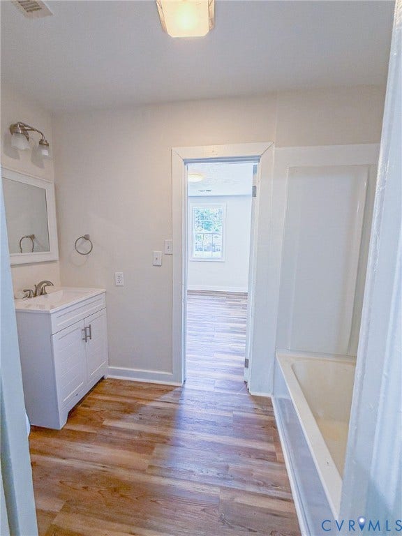 Wood-finish flooring, white vanity with integrated sink, wall-mounted mirror, and a built-in bathtub