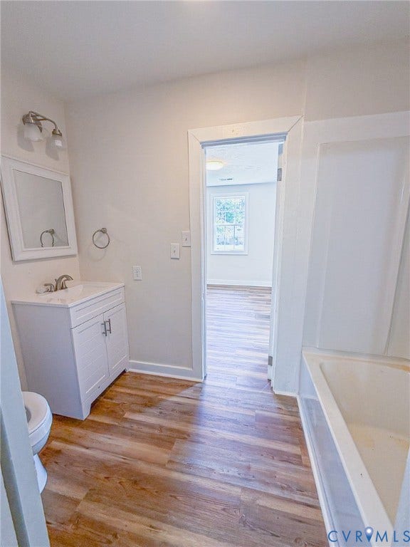 Bathroom featuring wood-finish flooring, a white vanity with integrated sink, a wall-mounted mirror, and a built-in tub with white surround