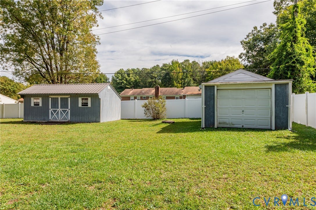 Expansive grassy area featuring a detached garage with a white roll-up door, a separate storage shed with a metal roof, and a white privacy fence