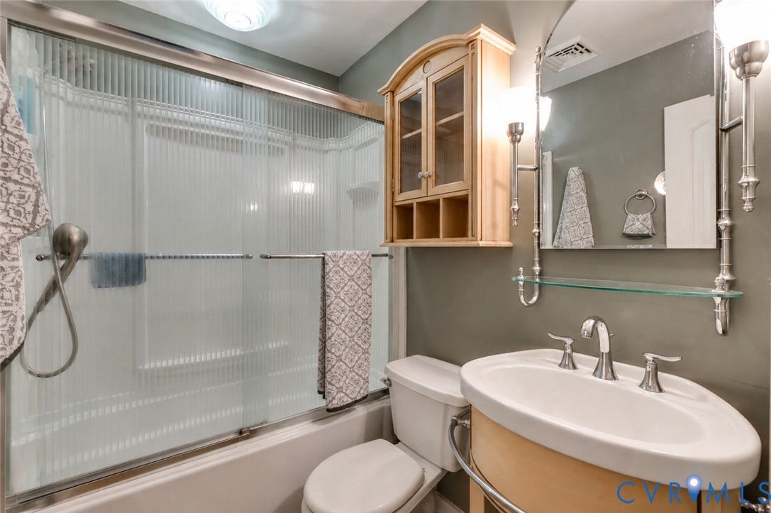 Tub/shower combination with sliding glass doors, single vanity with oval sink, wall-mounted mirror, and wood-finish wall cabinet