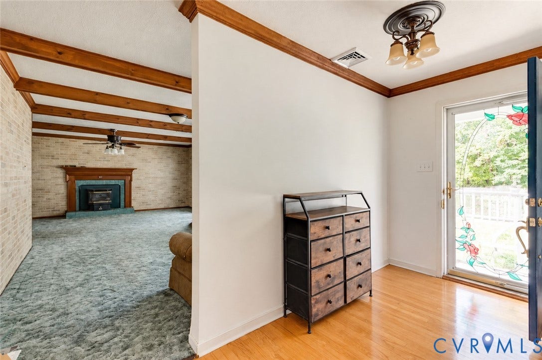 Hardwood flooring, decorative glass paneled door, fireplace with wood mantel, exposed wood ceiling beams, and brick accent wall