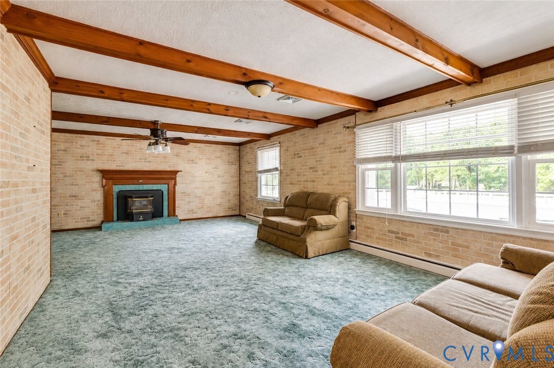 Exposed beam ceiling, brick accent walls, wood-mantled fireplace with a hearth, ceiling fan, and multiple windows