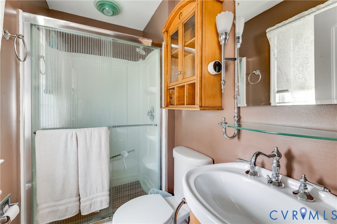 Shower with sliding glass enclosure, pedestal sink with chrome faucet, wall-mounted mirror with integrated lighting, and a built-in wooden cabinet