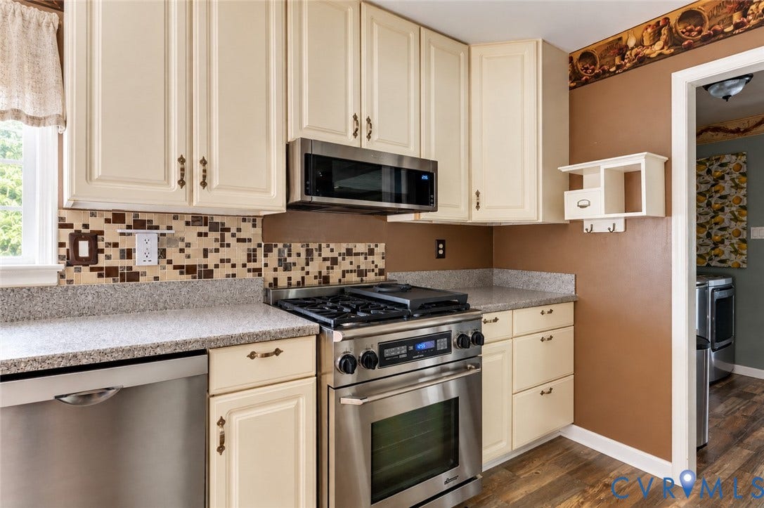 Cream-painted cabinetry, mosaic tile backsplash, stainless steel appliances, speckled countertops, and wood-finish flooring