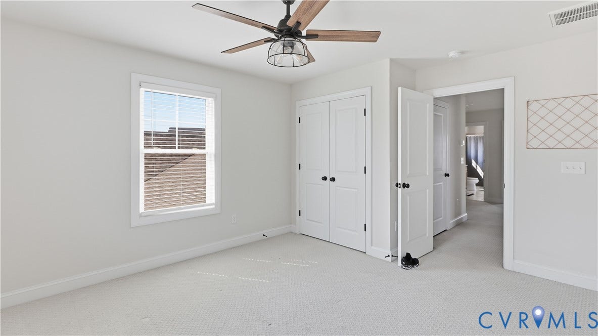 Bright primary bedroom featuring neutral carpet flooring, ceiling fan with integrated lighting, and multiple closet options including double doors for convenient storage access