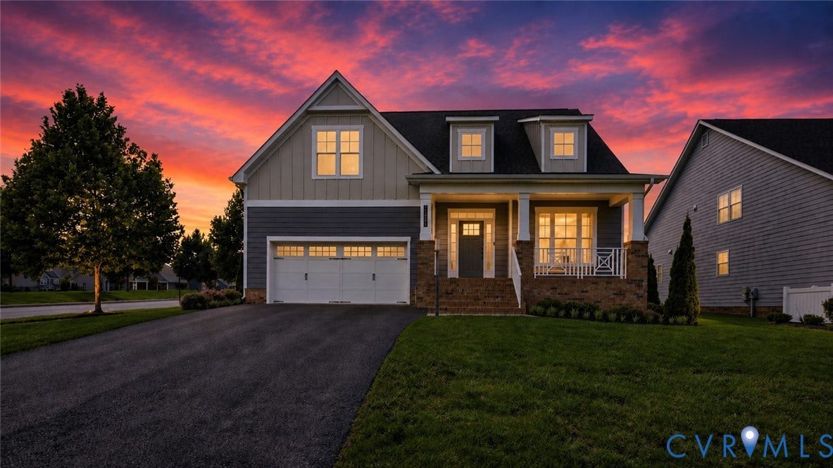 Two-car garage, dormer windows, covered front porch with white railings, brick foundation, and dark siding with light trim