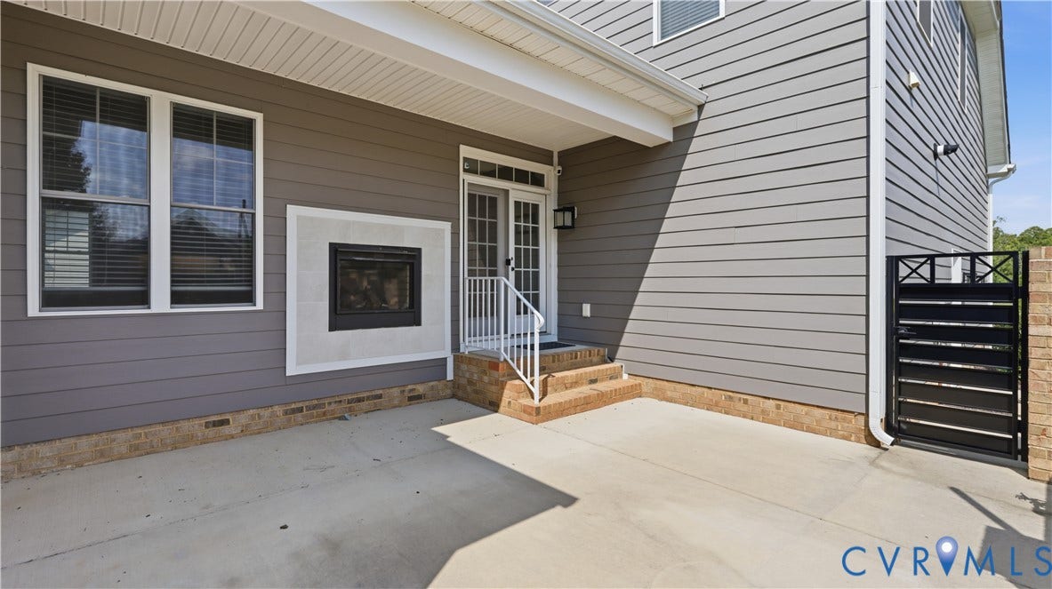 Outdoor patio with a built-in fireplace, concrete flooring, brick foundation, and a black wrought iron gate