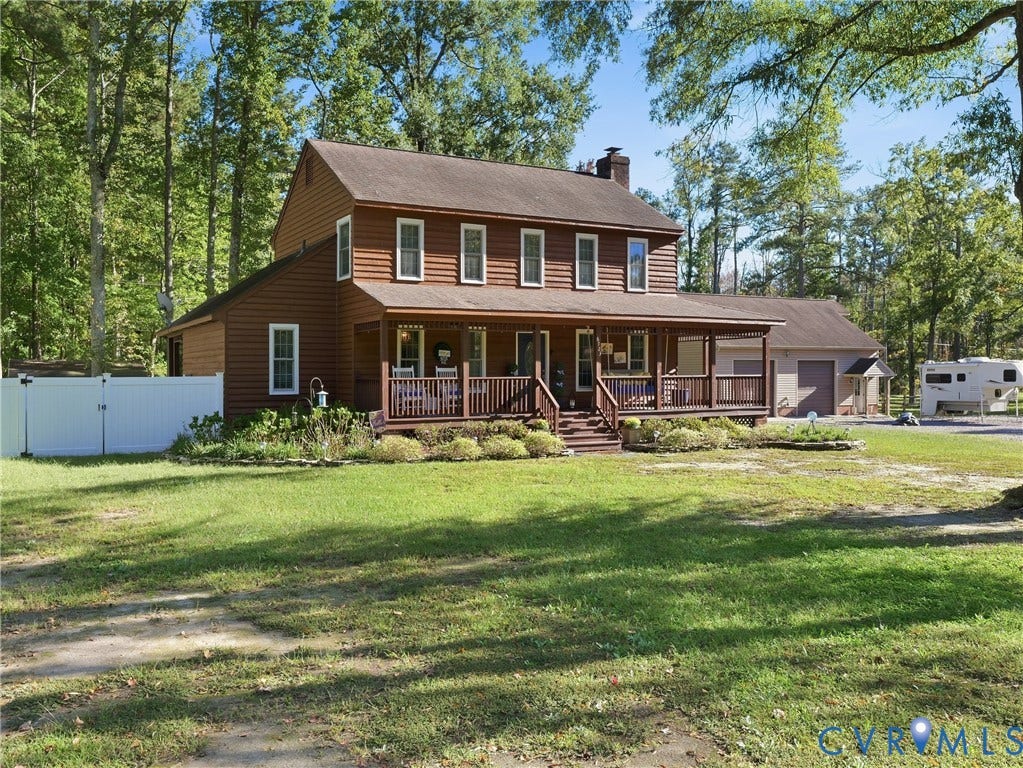 6723 Bull Hill Road, Prince George, VA 23875 Two-story home with horizontal siding, a wrap-around porch featuring wood railing, a brick chimney, and a detached two-car garage, all situated on a wooded lot
