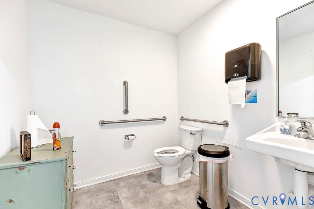 White wall finishes, toilet with grab bars, wall-mounted sink with faucet, large mirror, and gray flooring
