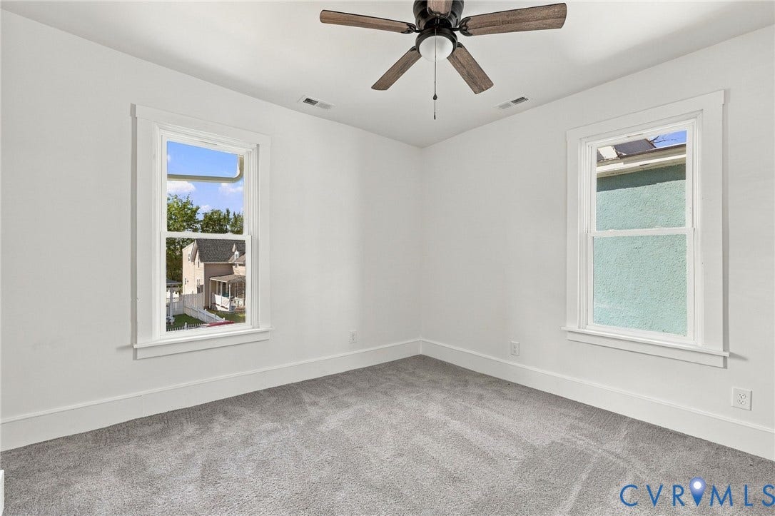 Interior space featuring neutral wall tones, gray carpeting, dual double-hung windows with white trim, and a ceiling-mounted fan with wooden blades