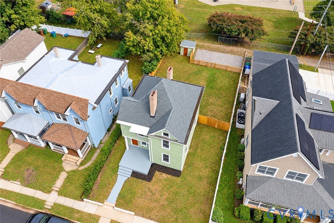Aerial view of a two-story home featuring light green siding, a gable roof with a central brick chimney, a concrete front porch, and a fenced backyard with a storage structure