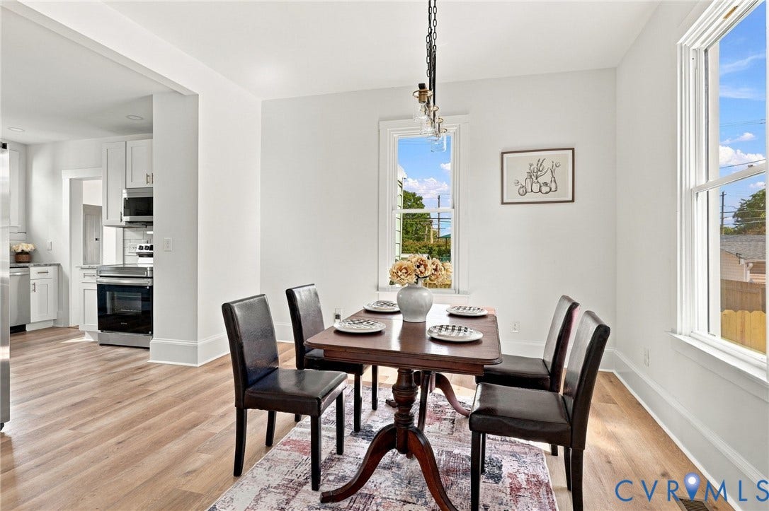 Open-concept dining area featuring wood-finish flooring, white walls, double-hung windows, and a multi-bulb hanging pendant light fixture