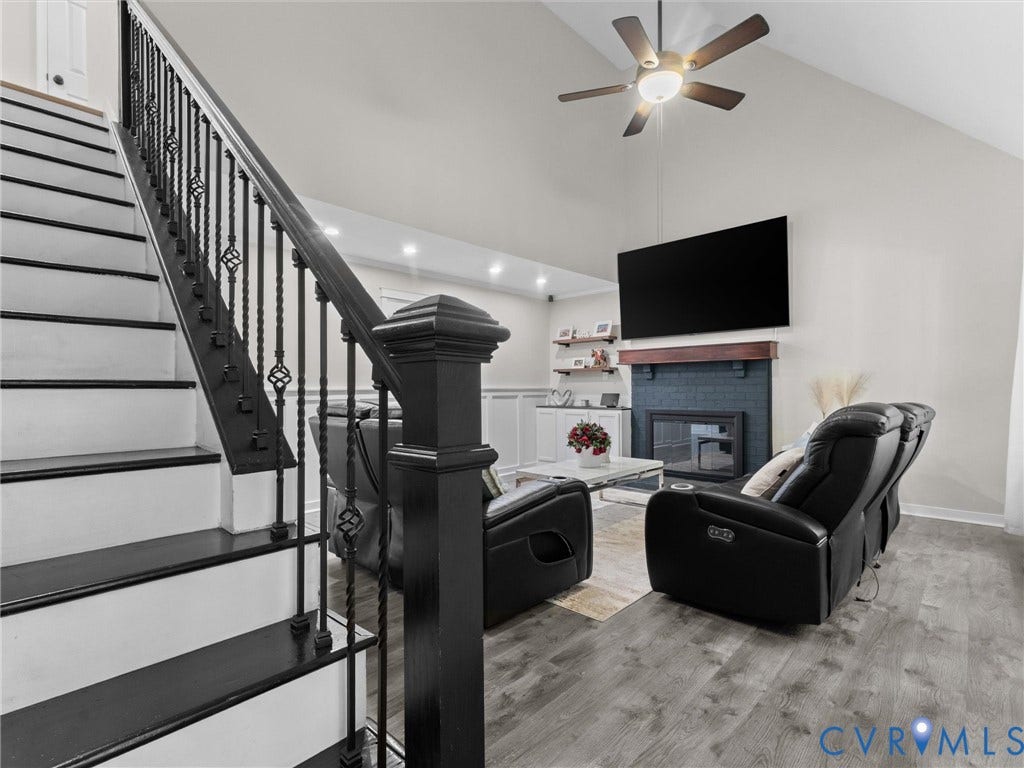 Vaulted ceiling with ceiling fan, black painted fireplace with wood mantel, built-in shelving, black wrought iron stair railing, and wood-finish flooring