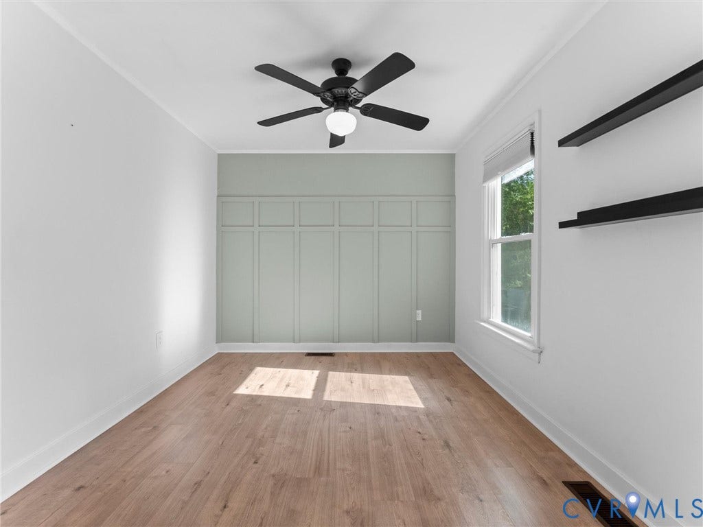 Wood-finish flooring, accent wall with panel detailing, ceiling fan with integrated lighting, built-in shelving, and a single window