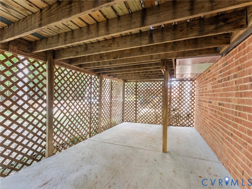 Covered patio area with a concrete surface, lattice paneling, exposed wood ceiling beams, and brick wall