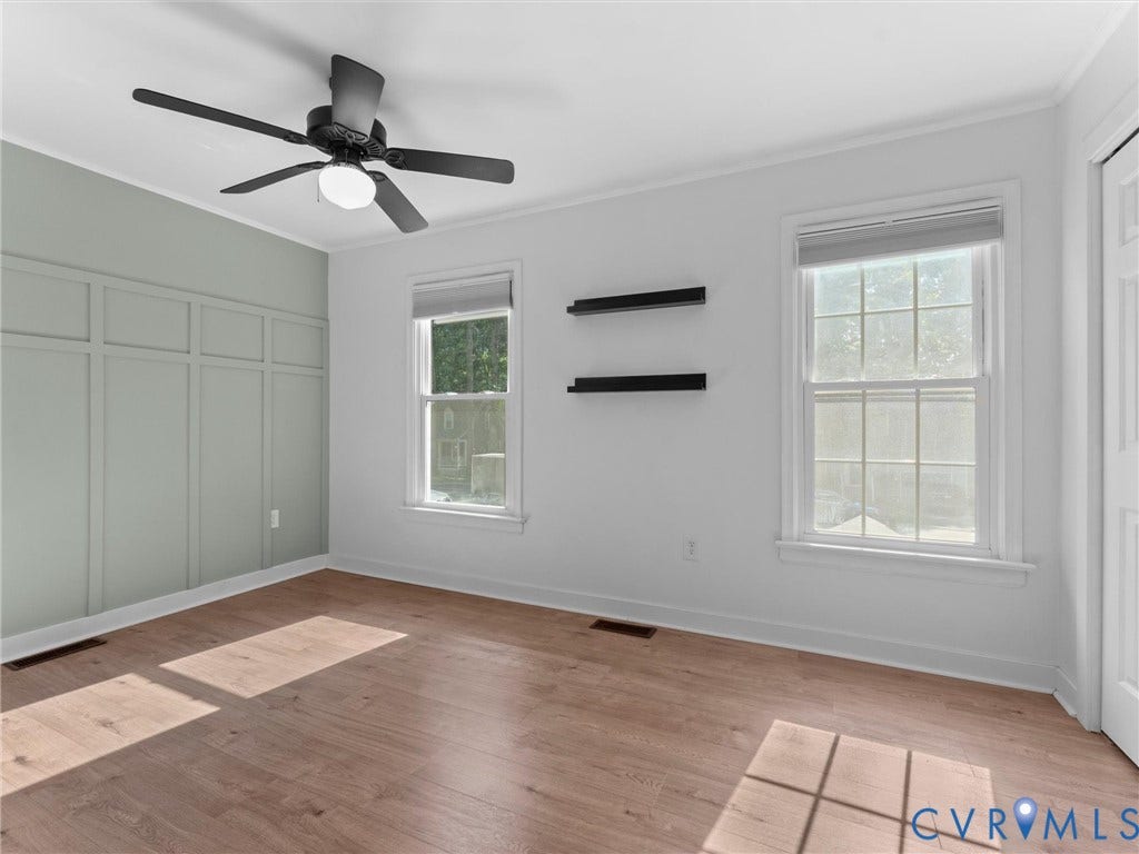 Wood-finish flooring, accent wall with wainscoting, two double-hung windows, ceiling fan with integrated lighting, and crown molding