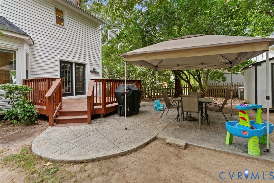 Rear exterior featuring white horizontal siding, a red-stained wood deck with railings, black-framed glass patio doors, and a patterned concrete patio