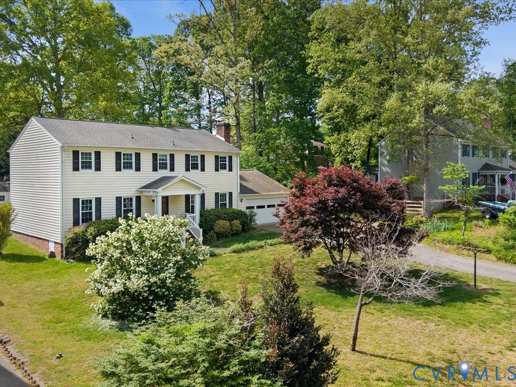 Colonial-style residence featuring yellow siding, a covered porch entry, black louvered shutters, an attached garage, and mature landscaping