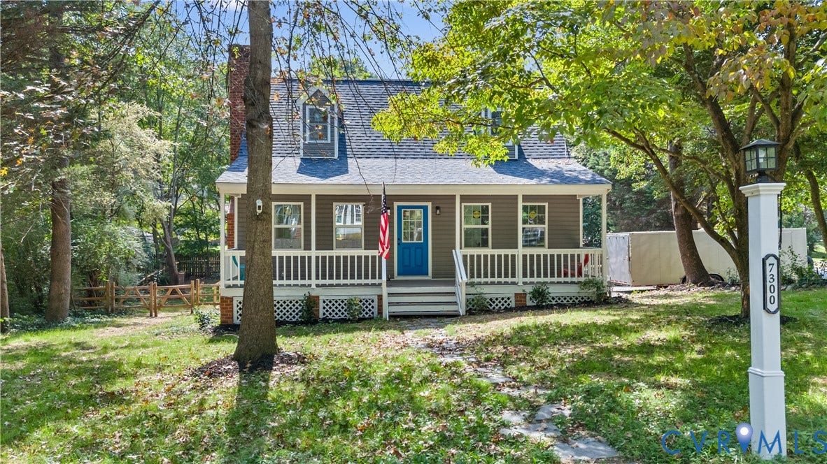 Cottage-style residence featuring a covered front porch with white railing, dormer windows, a brick chimney, and a post light with house numbers