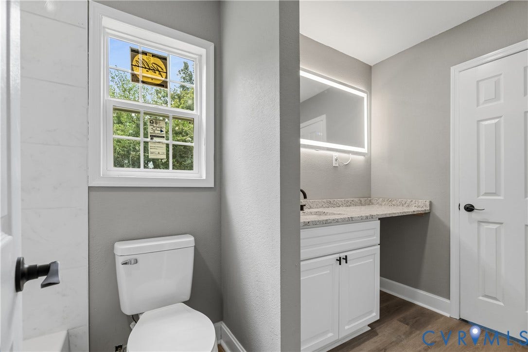 Bathroom featuring a window, white vanity with granite-style countertop, integrated lighting mirror, and wood-finish flooring