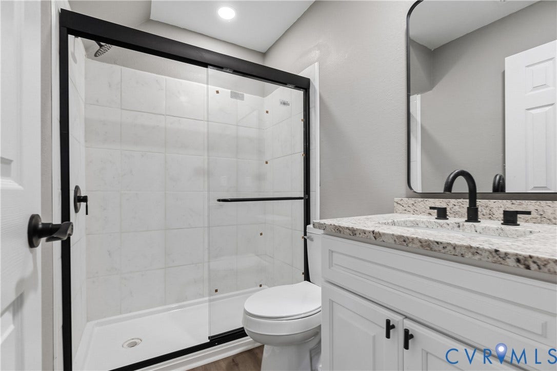 White panel cabinetry with black hardware, stone-finish countertop, black curved faucet, frameless mirror, and glass-enclosed shower with white tile surround