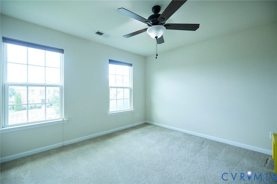 Neutral tone wall paint, carpeted flooring, two windows with blinds, and a ceiling fan with integrated lighting