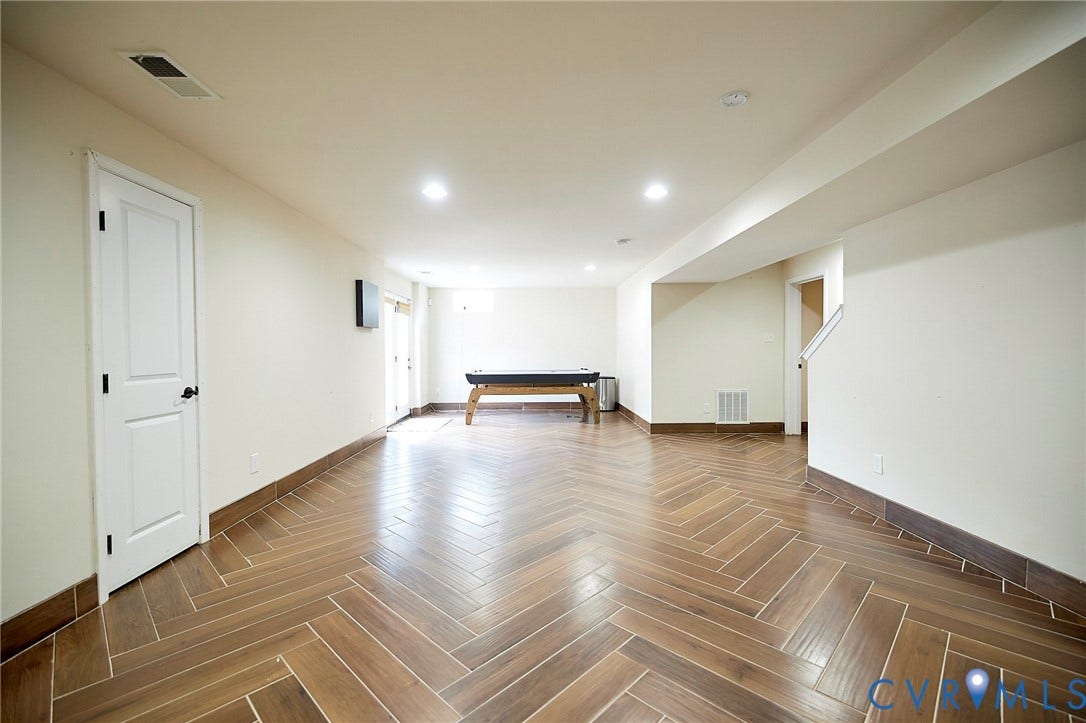 Spacious room featuring wood-finish flooring with a herringbone pattern, recessed lighting, white interior doors, and a white staircase with a half-wall