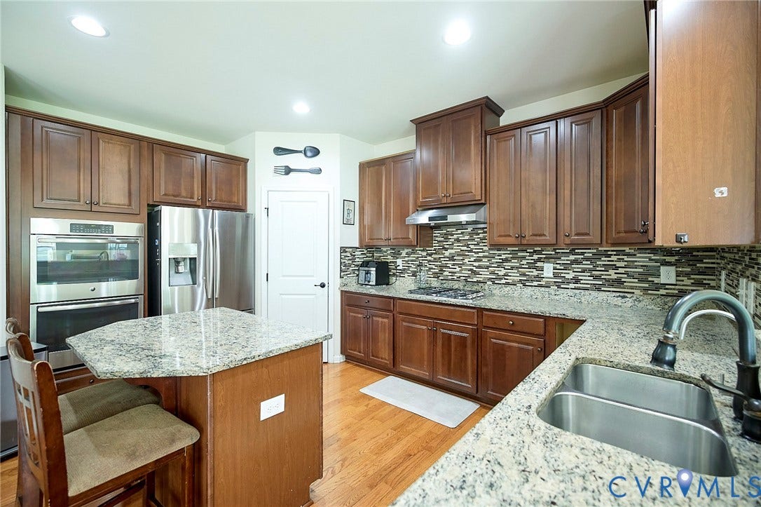 Kitchen featuring wood-finish cabinetry, stainless steel appliances, granite countertops, a decorative tile backsplash, and a double basin sink