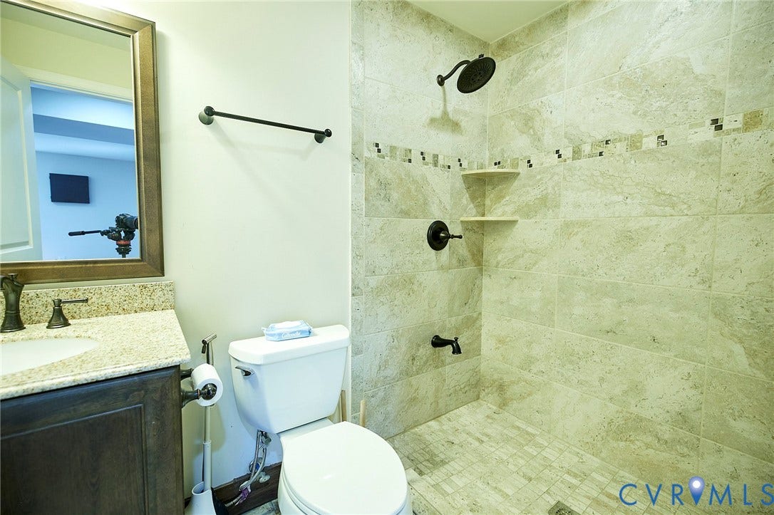 Granite-finish countertop with an undermount sink, bronze-finish fixtures, a tiled shower with corner shelving, and a decorative tile accent band