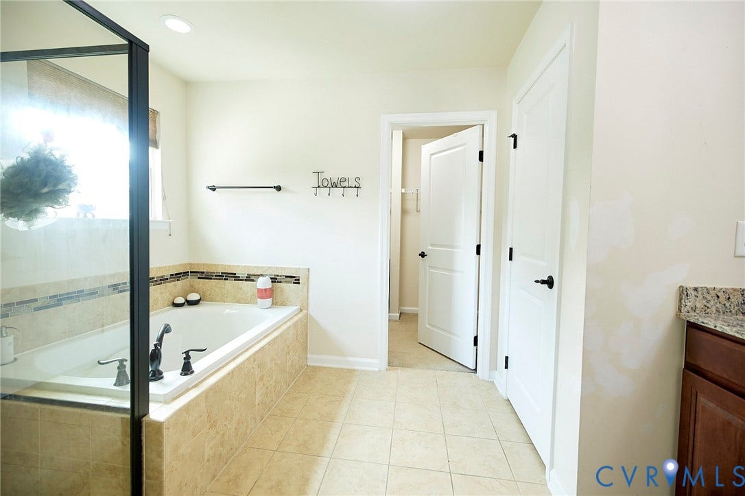 Tiled soaking tub with dark bronze fixtures, glass-enclosed shower, neutral wall paint, and a door with dark hardware