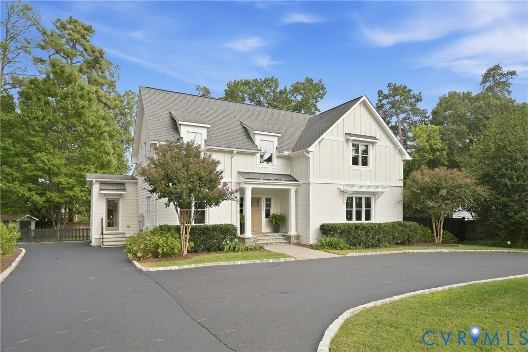 White exterior with board and batten siding, dark shingle roof, paved driveway, mature trees, and established landscaping