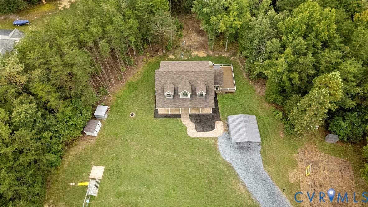 Gabled roofline with dormer windows, extended rear deck, gravel driveway, covered carport, and multiple storage sheds