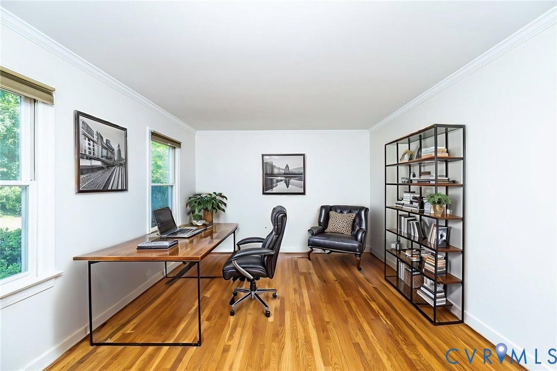Bright home office featuring hardwood flooring, large windows with natural light, a desk with metal frame and wood top, ergonomic office chair, and built-in shelving unit with geometric grid design