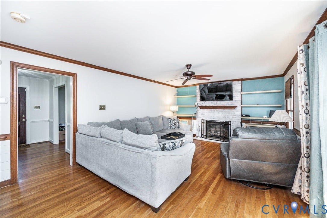 Spacious living room featuring hardwood flooring, a wood-burning fireplace with built-in cabinetry and shelving, ceiling fan, and abundant natural light from large windows with custom treatments
