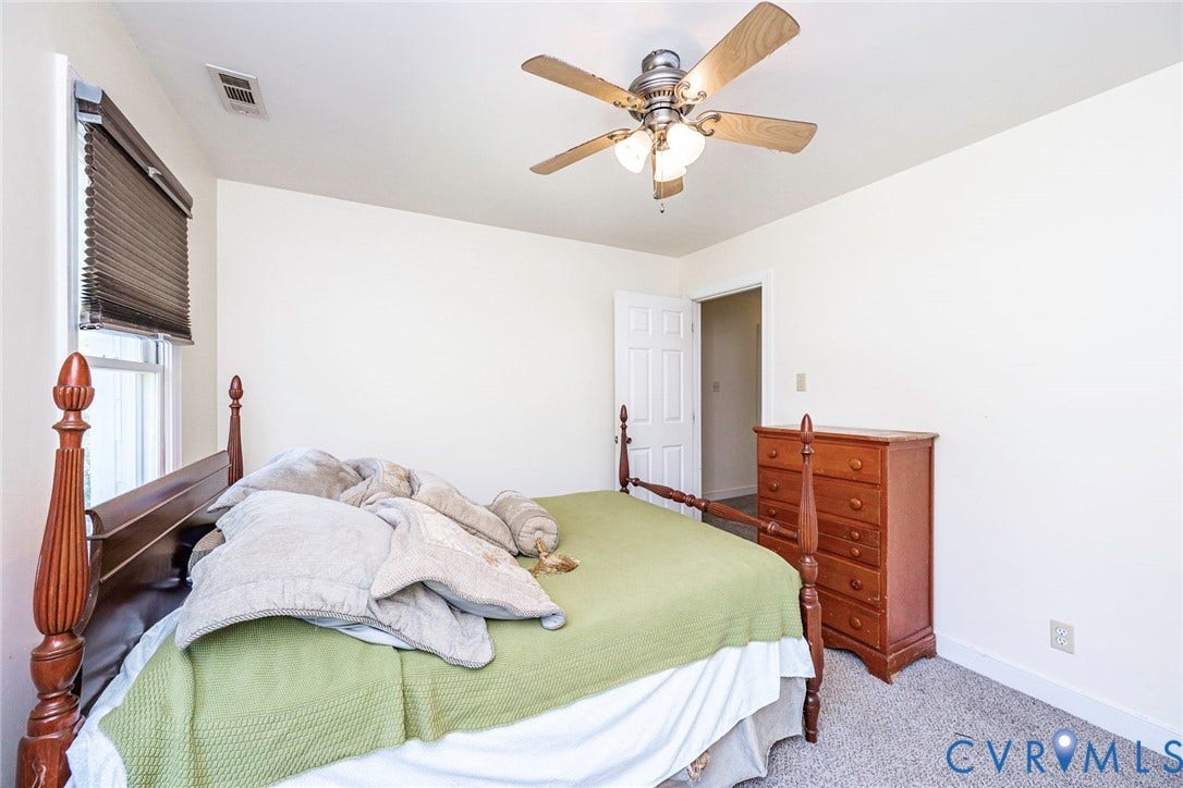 Spacious bedroom featuring hardwood flooring, carpet flooring, wooden post bed frame, wood-finish dresser with multiple drawers, ceiling fan with integrated lighting, and abundant natural light from window with roller shade