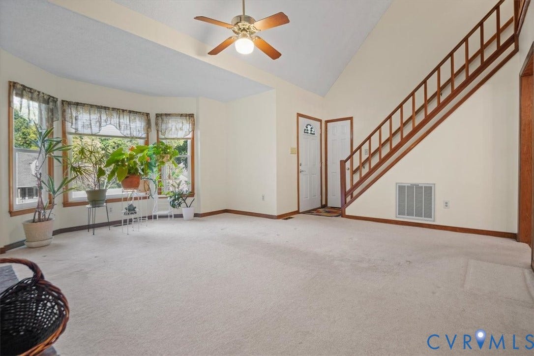 Vaulted ceiling with ceiling fan, bay window with wood trim, light-colored carpeting, and a wooden staircase