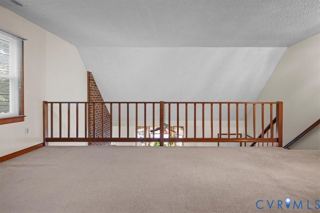 Carpeted loft area with wood balustrade, sloped ceiling, and brick architectural detail