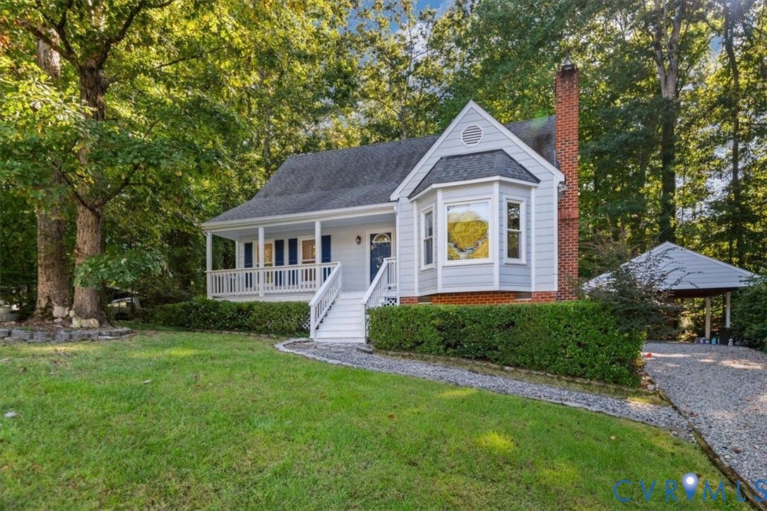 Light blue siding exterior with a dark roof, a covered porch with white railings, a prominent bay window, and an attached brick chimney
