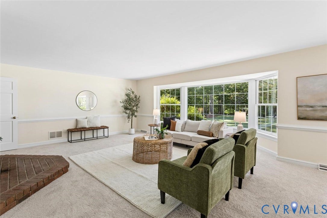 Expansive living area featuring a large bay window, light-toned carpeting, white trim, and a brick hearth