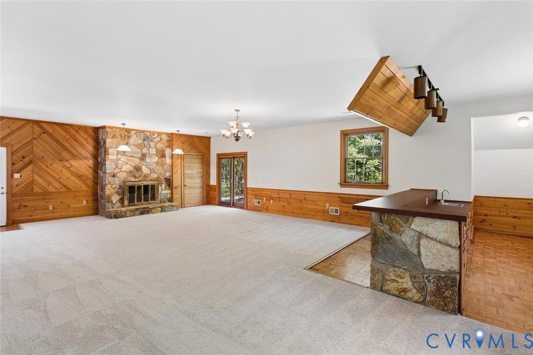 Stone fireplace with metal grate, wood-paneled accent wall, built-in wet bar with stone base, glass-paned double doors, and chandelier lighting