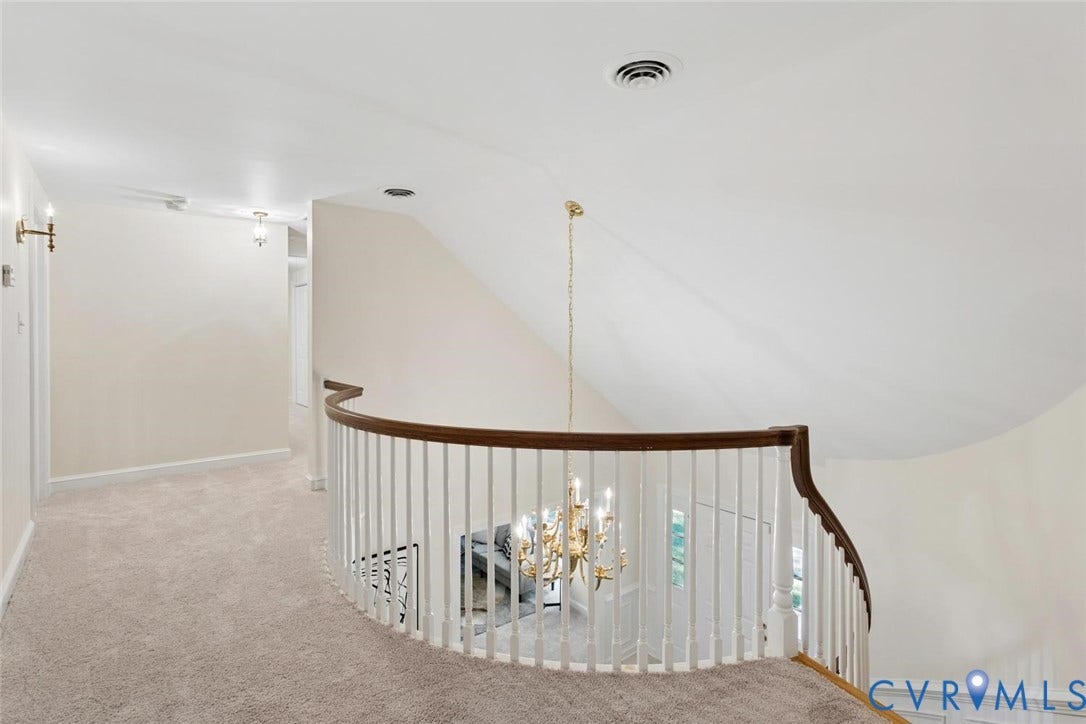 Carpeted hallway featuring a curved railing with wood-finish top rail, white balusters, and a brass chandelier