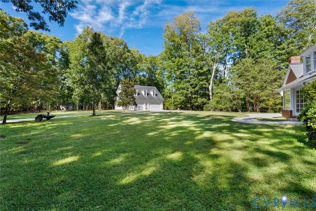 Expansive lawn bordered by mature trees, featuring a detached structure with dormer windows and a paved driveway